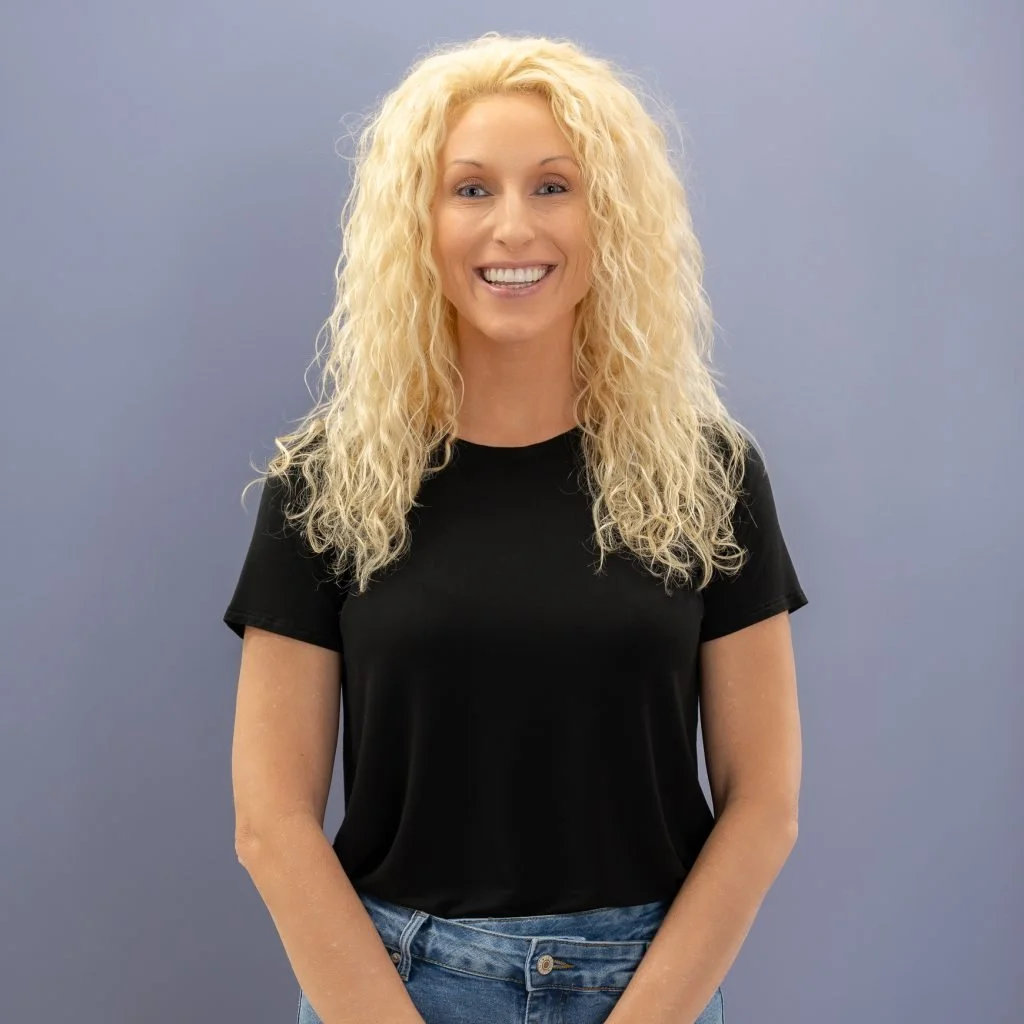 A woman with long, curly blonde hair smiling at the camera, wearing a black t-shirt and blue jeans against a plain light blue background.