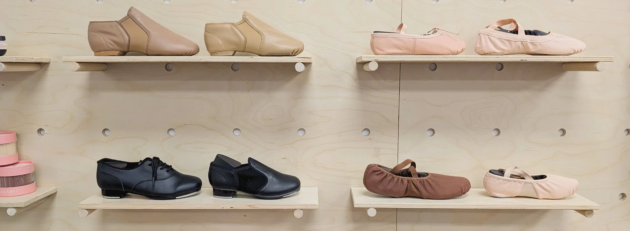 Display of ballet shoes and tap shoes on shelves against a light wood wall.