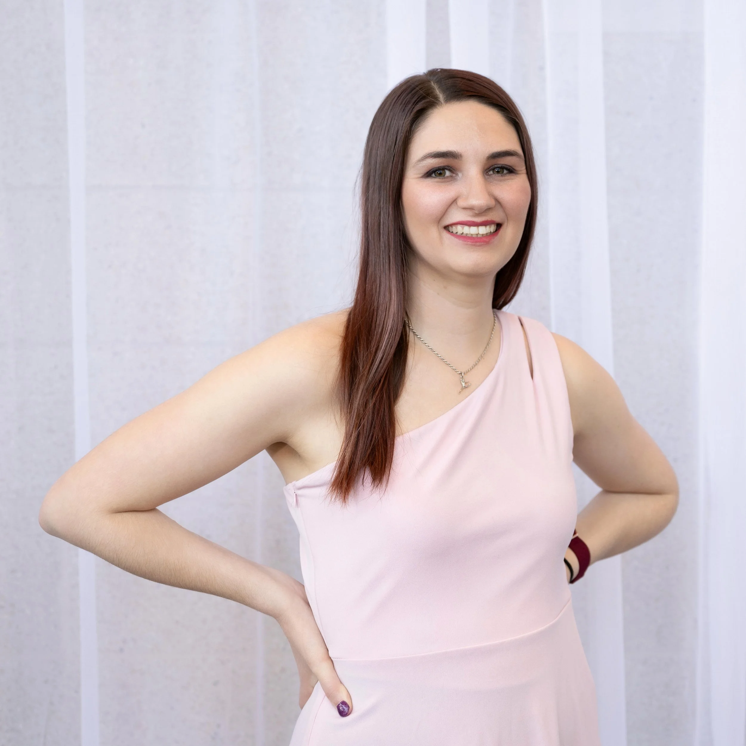 A woman with long brown hair and light skin, wearing a pink sleeveless top, smiling at the camera, with her right hand resting on her hip, against a light gray tiled wall background.
