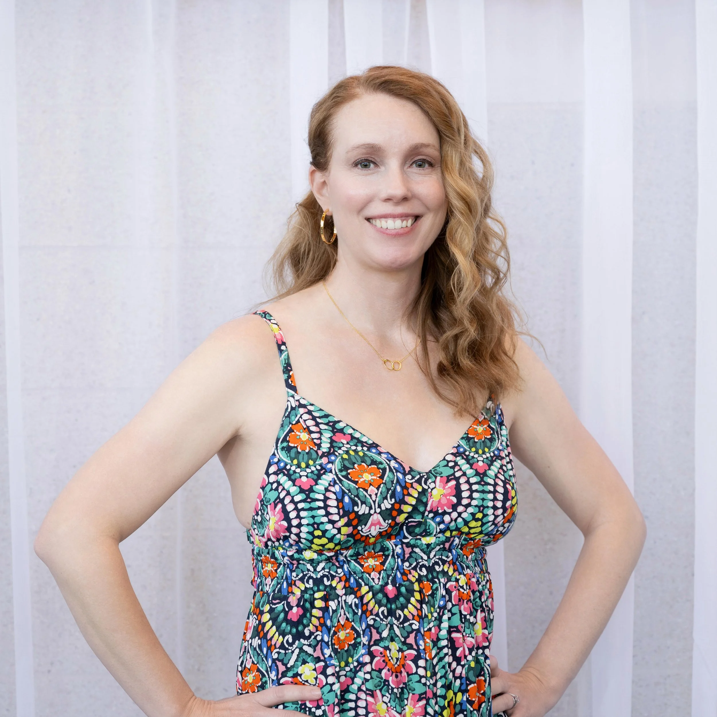 A woman with curly red hair wearing a colorful floral dress and gold jewelry, smiling with her hand on her hip in front of a light-colored textured wall.