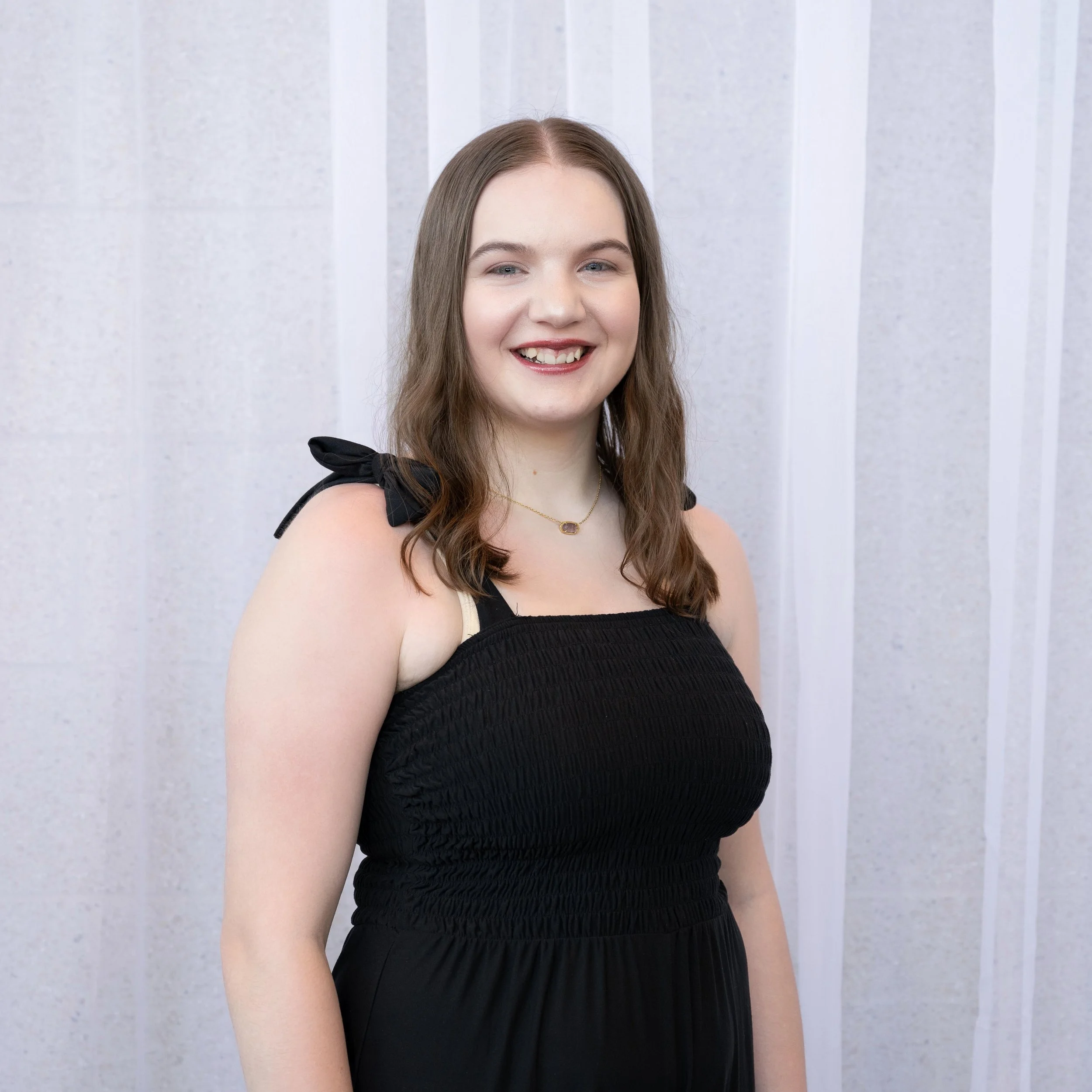 A woman with brown hair smiling, wearing a black sleeveless top with shoulder bows, standing in front of a light-colored, vertically striped background.