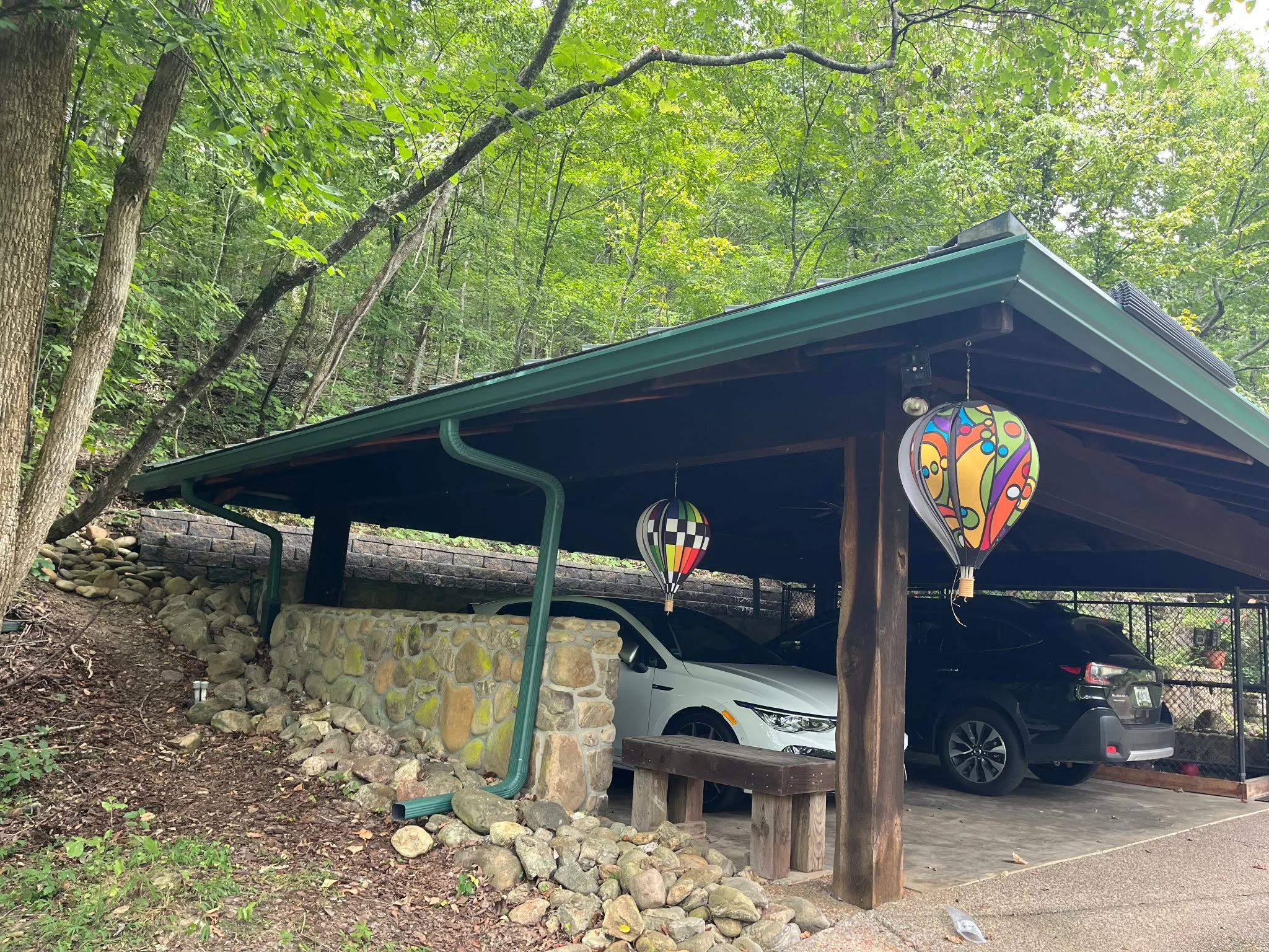 Parking area with cars parked under a shelter with a green metal roof, surrounded by trees and greenery, with decorative lanterns hanging from the shelter.