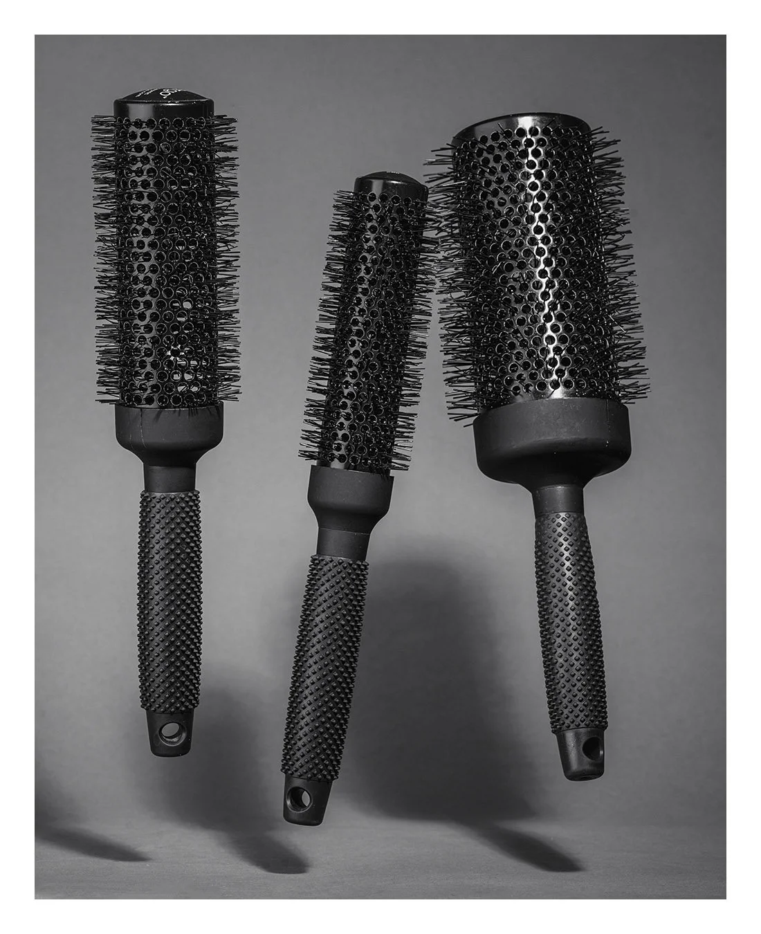 Three black hairbrushes with textured handles and rounded barrels, arranged in a row on a plain gray background.