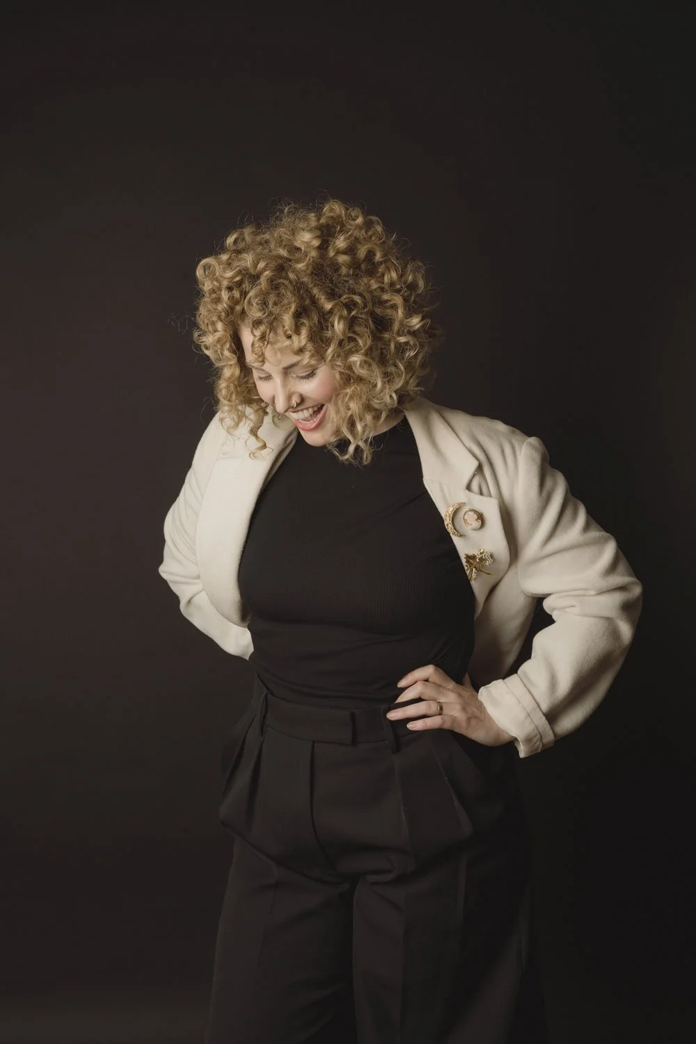 A woman with curly blonde hair wearing a black top, a white blazer with pins, and black pants, smiling against a dark background.