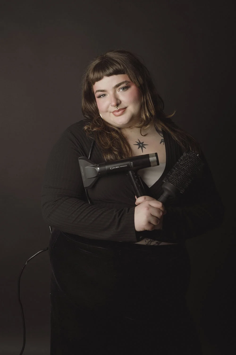 A woman with curly brown hair and tattoos holding a hairdryer and a microphone against a dark background.