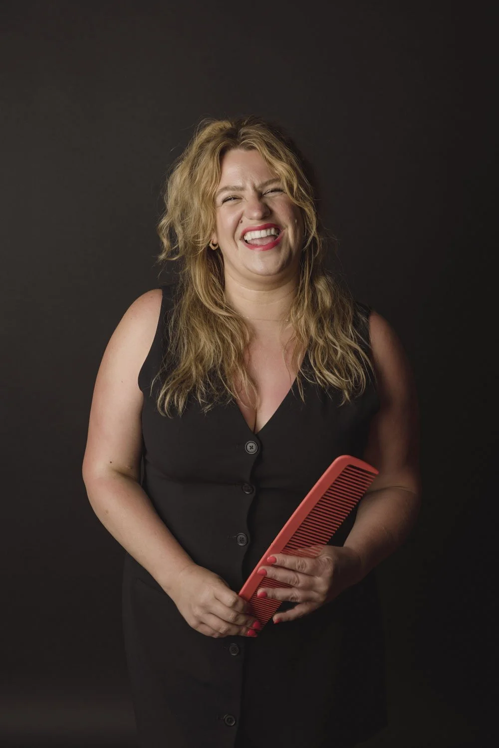 A woman with curly blonde hair wearing a black sleeveless dress, holding a red comb, and smiling with her mouth open.