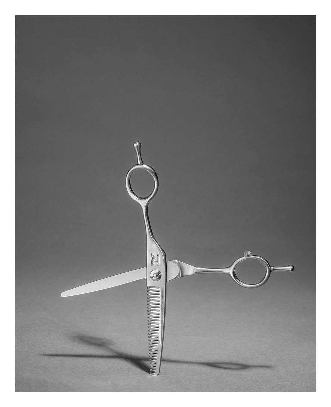 A pair of haircutting scissors with a blade and a measuring ruler handle, balanced on a gray surface with a plain gray background.