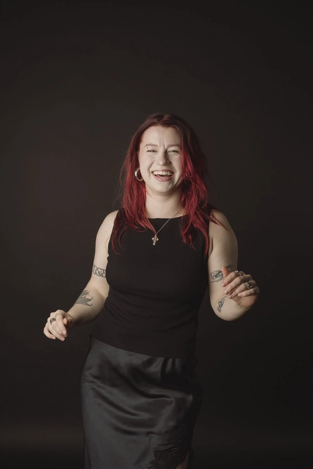 A young woman with red hair, wearing a black sleeveless top and satin skirt, laughing and dancing against a dark background.