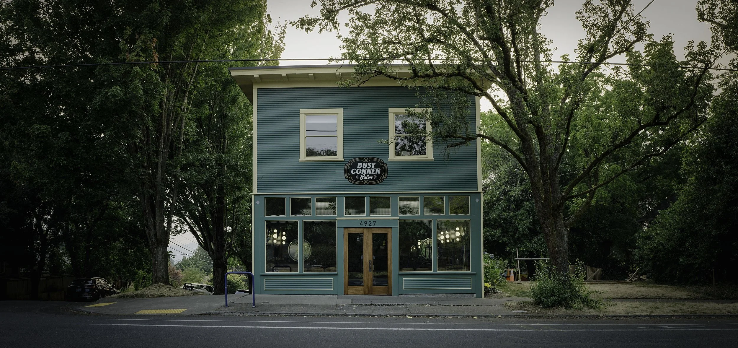 A two-story blue building with a salon named "Busy Corner Salon" on the sign, located at 4927. The building has large glass windows on the ground floor and two windows on the upper floor, surrounded by green trees and parked cars nearby.