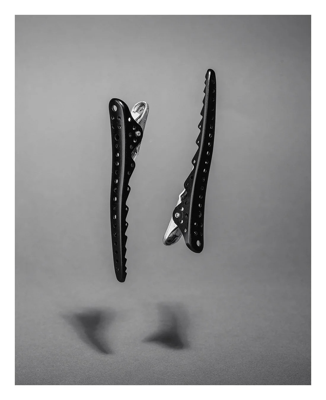 Two black hair clips with small holes, suspended in mid-air with shadows cast on gray background.