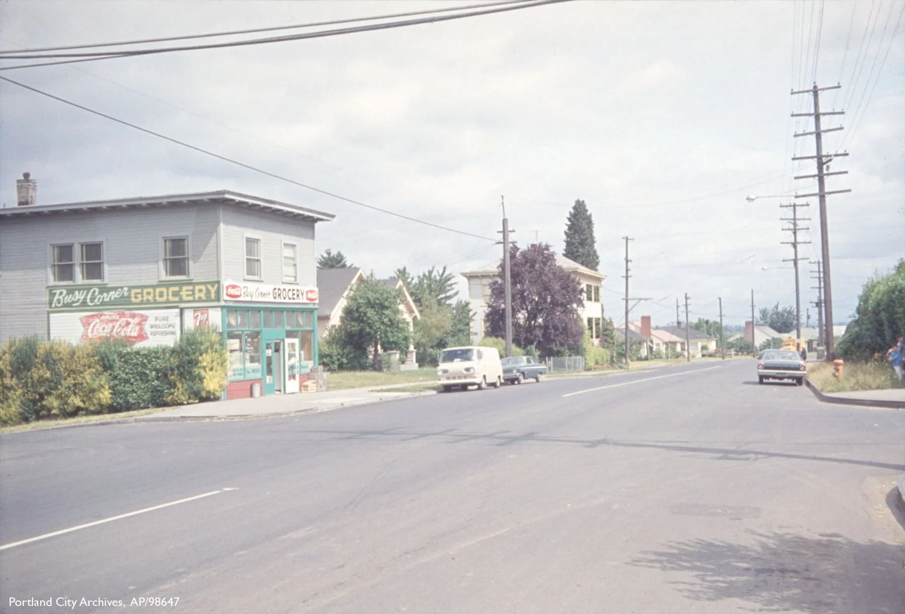 A small grocery store named Busy Corner Grocery on the corner of a street with residential houses, cars parked along the road, and utility poles with power lines. The store has a sign with Coca-Cola advertising and a teal-colored entrance.