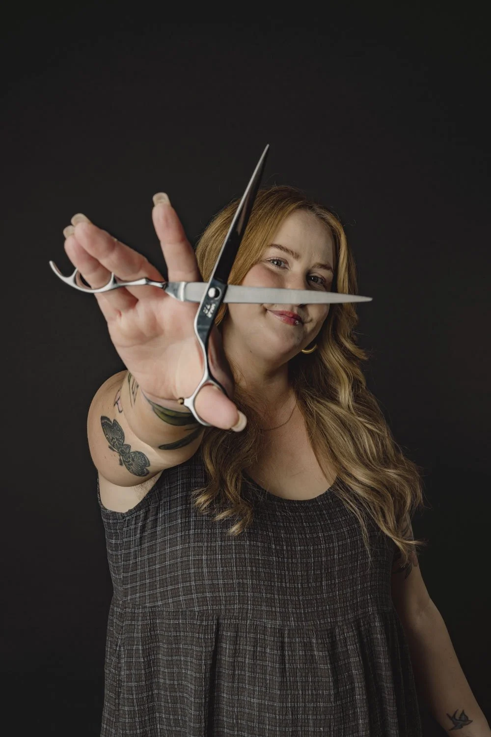 A woman with red hair and tattoos holding a pair of hair scissors toward the camera, smiling against a dark background.