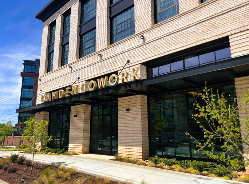 Exterior view of Camden Cowork, a modern building with large windows and a brick facade, with greenery and a clear blue sky.