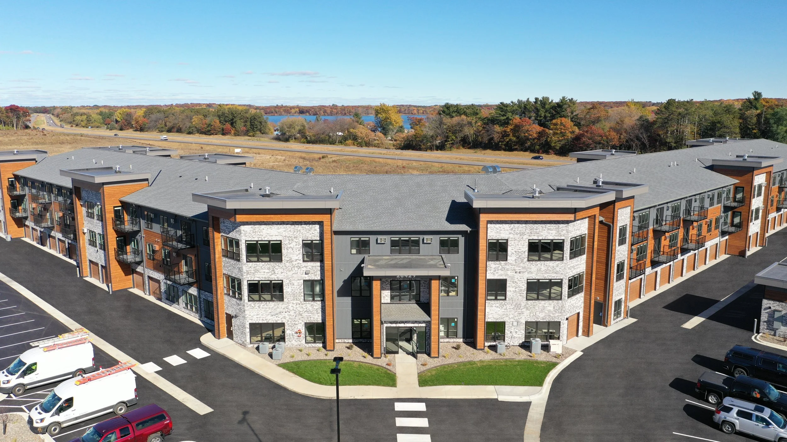 Aerial view of a modern multi-story apartment building with brick and stone exterior, surrounded by a parking lot with several vehicles, set against a background of trees, a highway, and a lake.