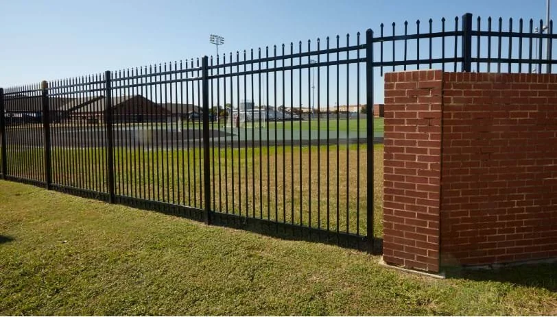 Black metal fence with brick pillar beside a grassy field and sports stadium in the background.