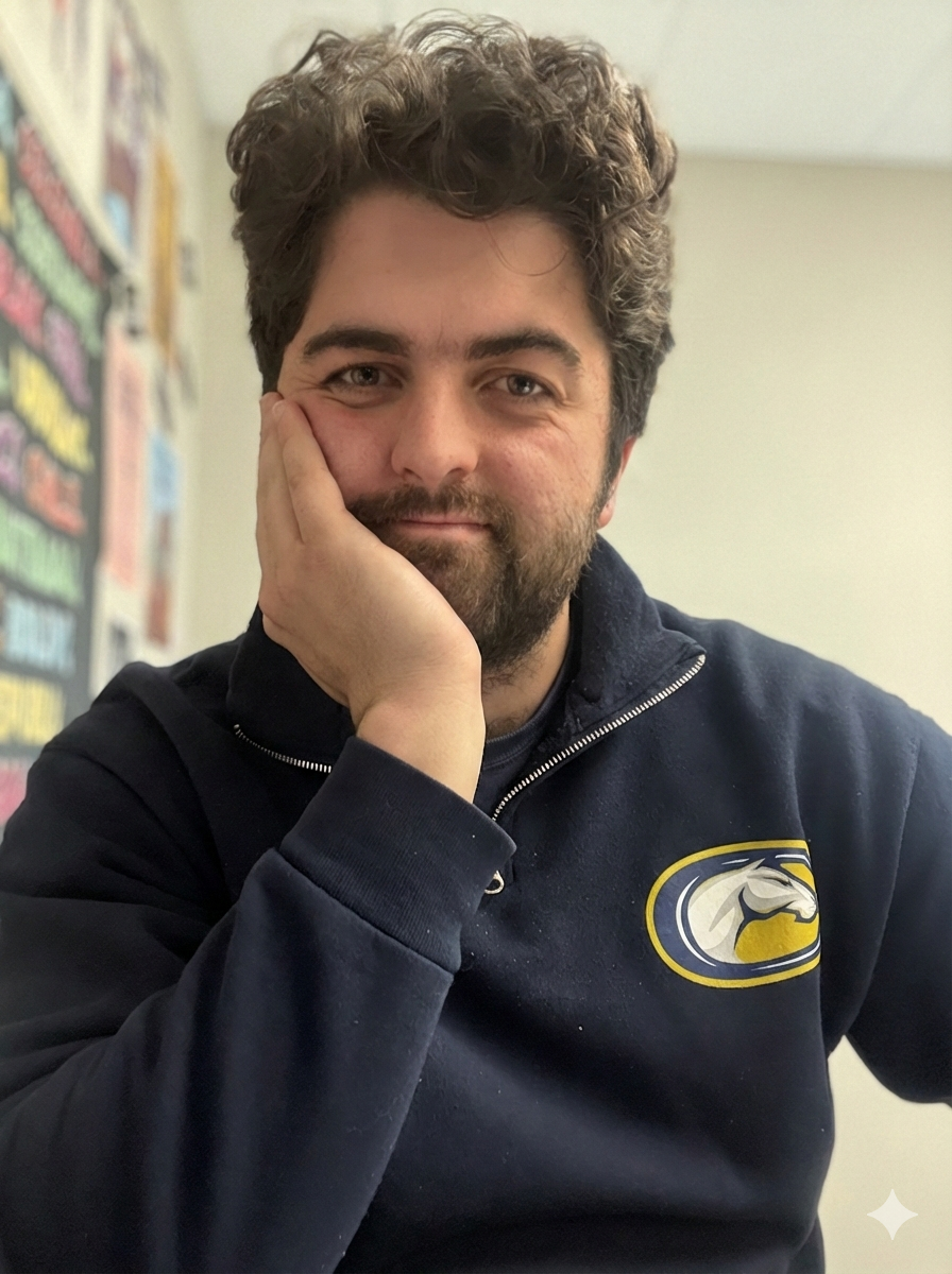 A man with curly brown hair and a beard, resting his face on his hand with a slight smile, wearing a black zip-up sweatshirt with a logo of a horse's head inside an oval patch on the chest, sitting in a room with a blurred bulletin board with colorful papers in the background.