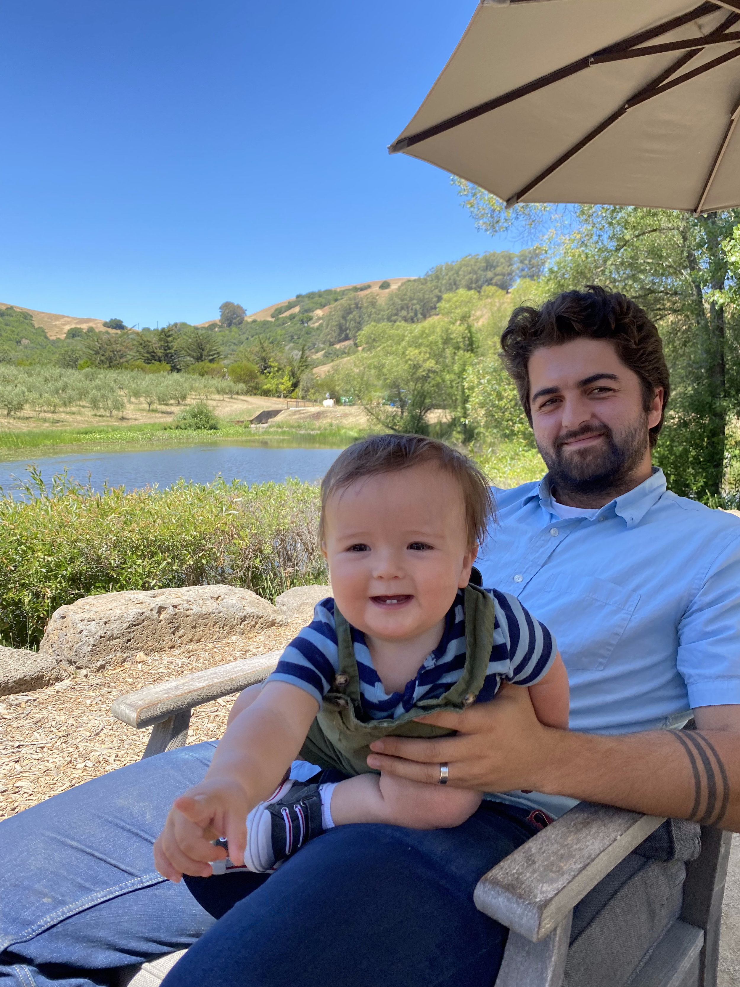 Harley Poston and his daughter on a park bench with a scenic background of rolling hills, trees, and a pond, under a blue sky with a large outdoor umbrella.