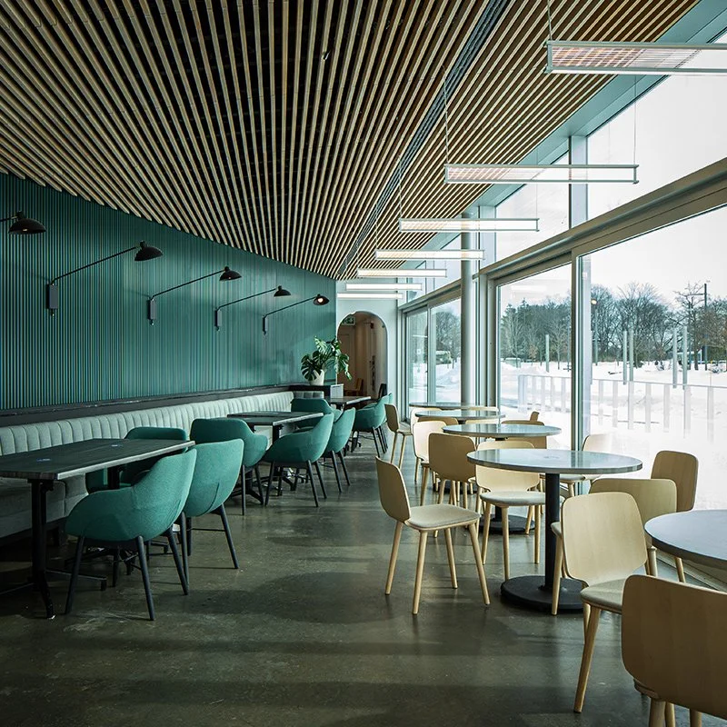 Intérieur d'un café moderne avec des tables en bois, des chaises en tissu vert et beige, une grande fenêtre laissant entrer la lumière naturelle, et un plafond en lattes de bois avec des luminaires suspendus.