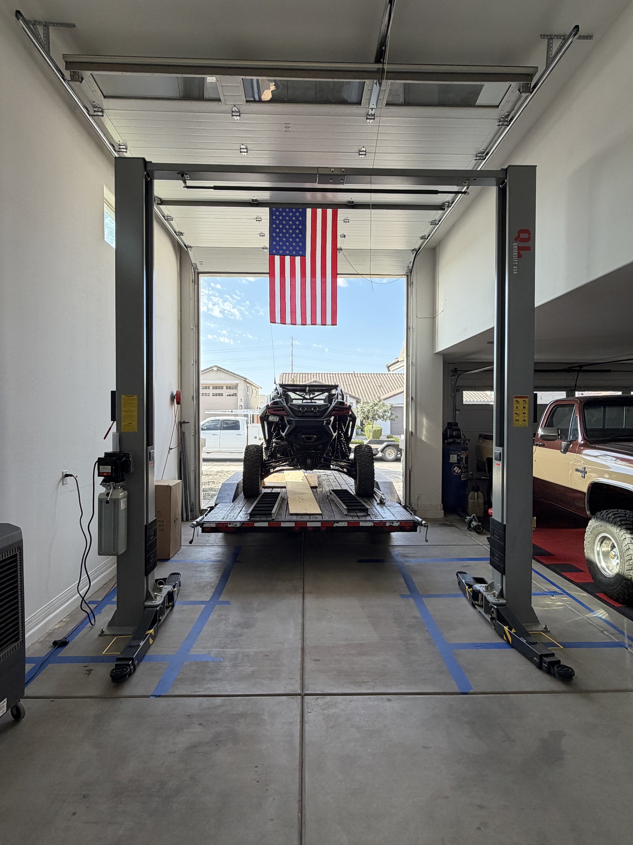 Garage with an American flag hanging from the ceiling, a vehicle on a lift, and a small truck parked inside.