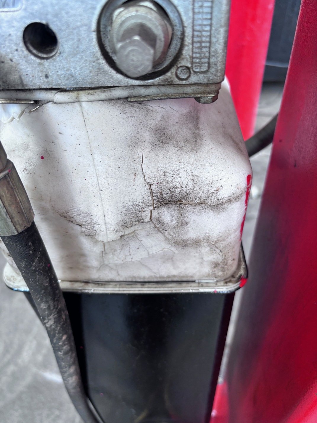 Close-up of a dirty, cracked white plastic container or casing with a metal bolt at the top, attached to a black surface. Part of a red frame is visible on the right side.
