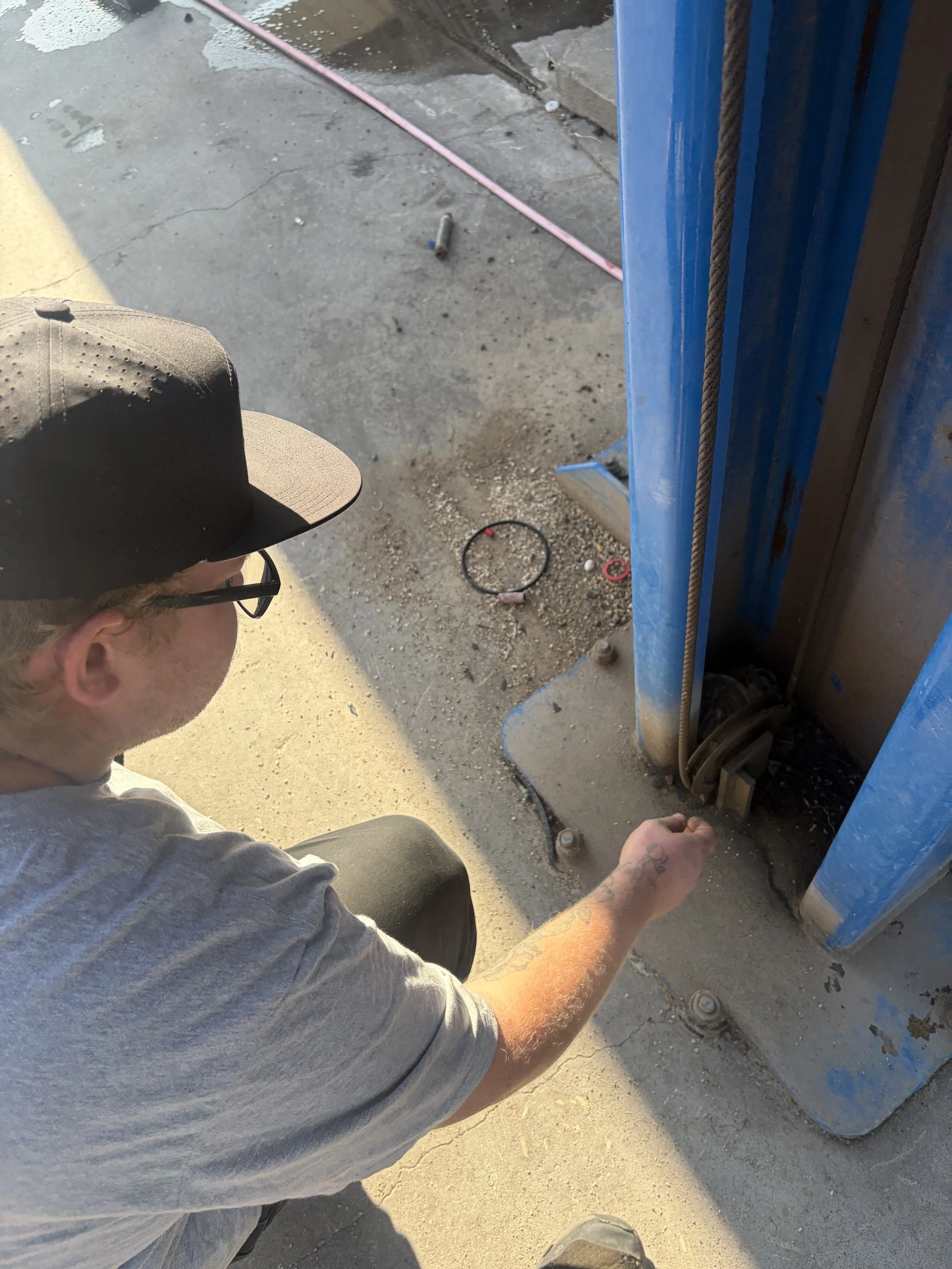 A man in a gray shirt with tattoos on his arm, wearing a black cap and glasses, is crouched down working near a blue hydraulic lift. The ground is concrete with dirt and small debris, and some tools and cables are visible around him.