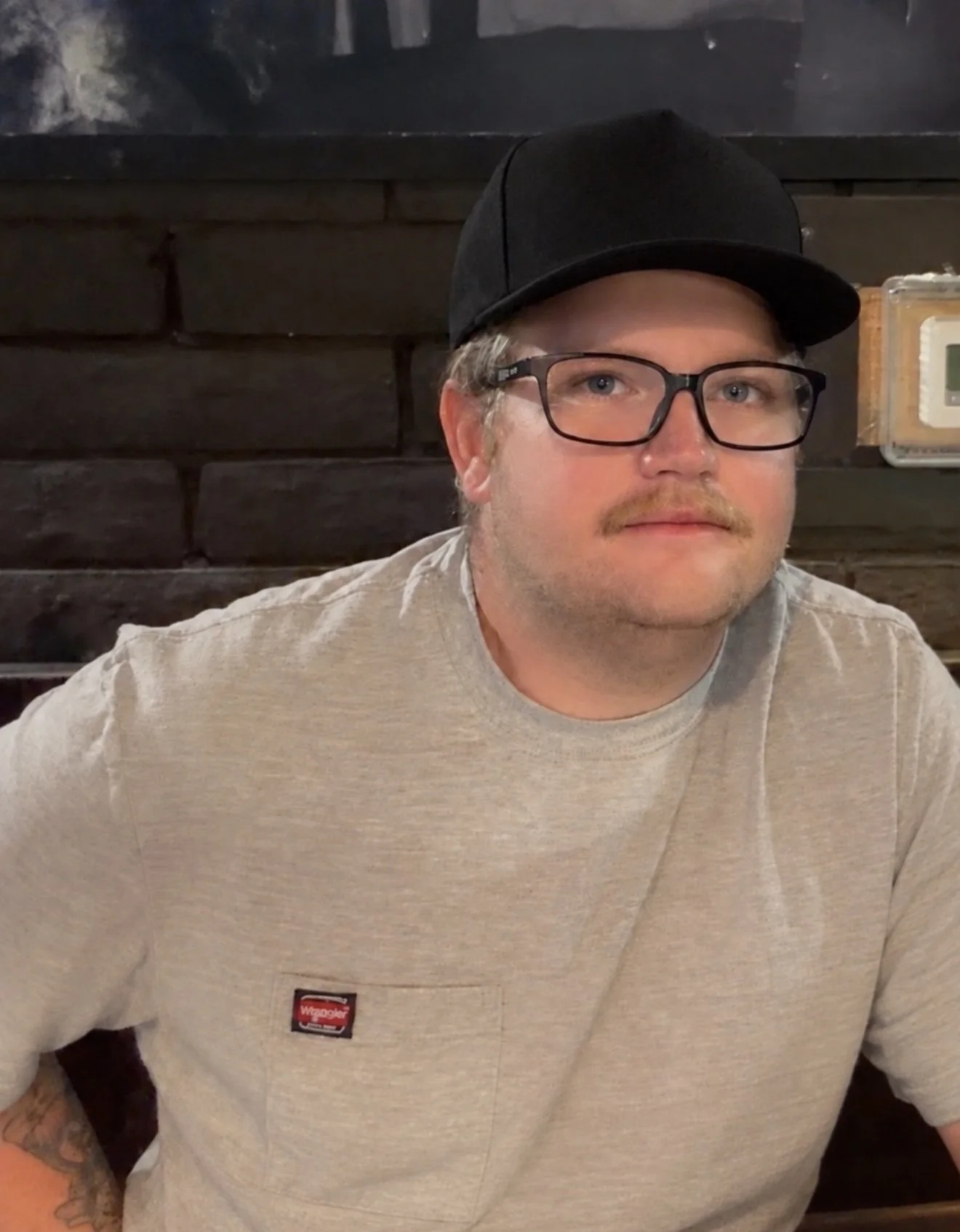A man wearing glasses, a black cap, and a beige T-shirt is sitting indoors against a black brick wall. The man has light hair, a mustache, and a hint of beard, and he is looking directly at the camera.