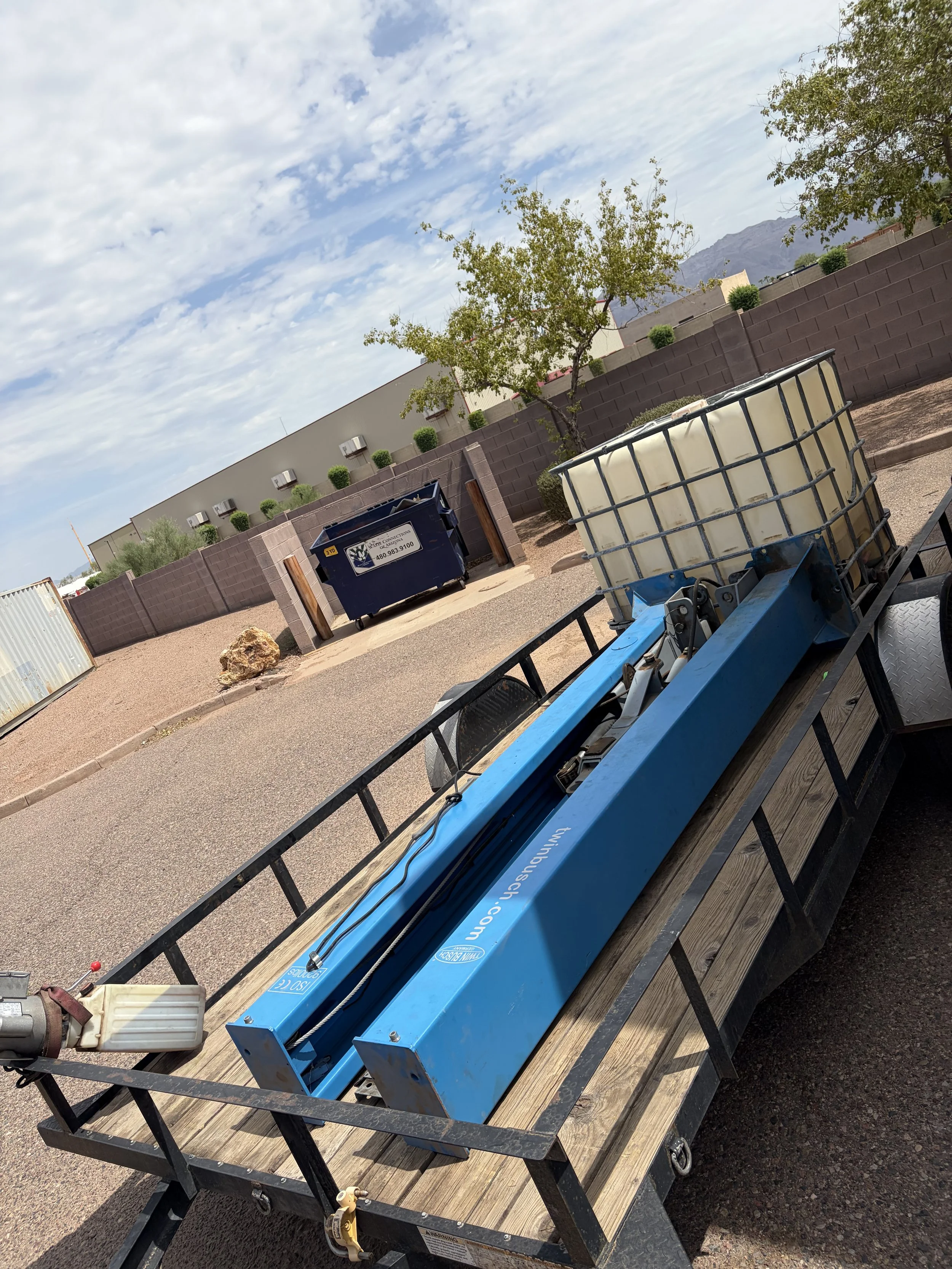 A flatbed cart with a blue industrial heat gun, a white plastic container, and a small box, parked outdoors on a paved surface next to a brown brick wall with trees and a building in the background.