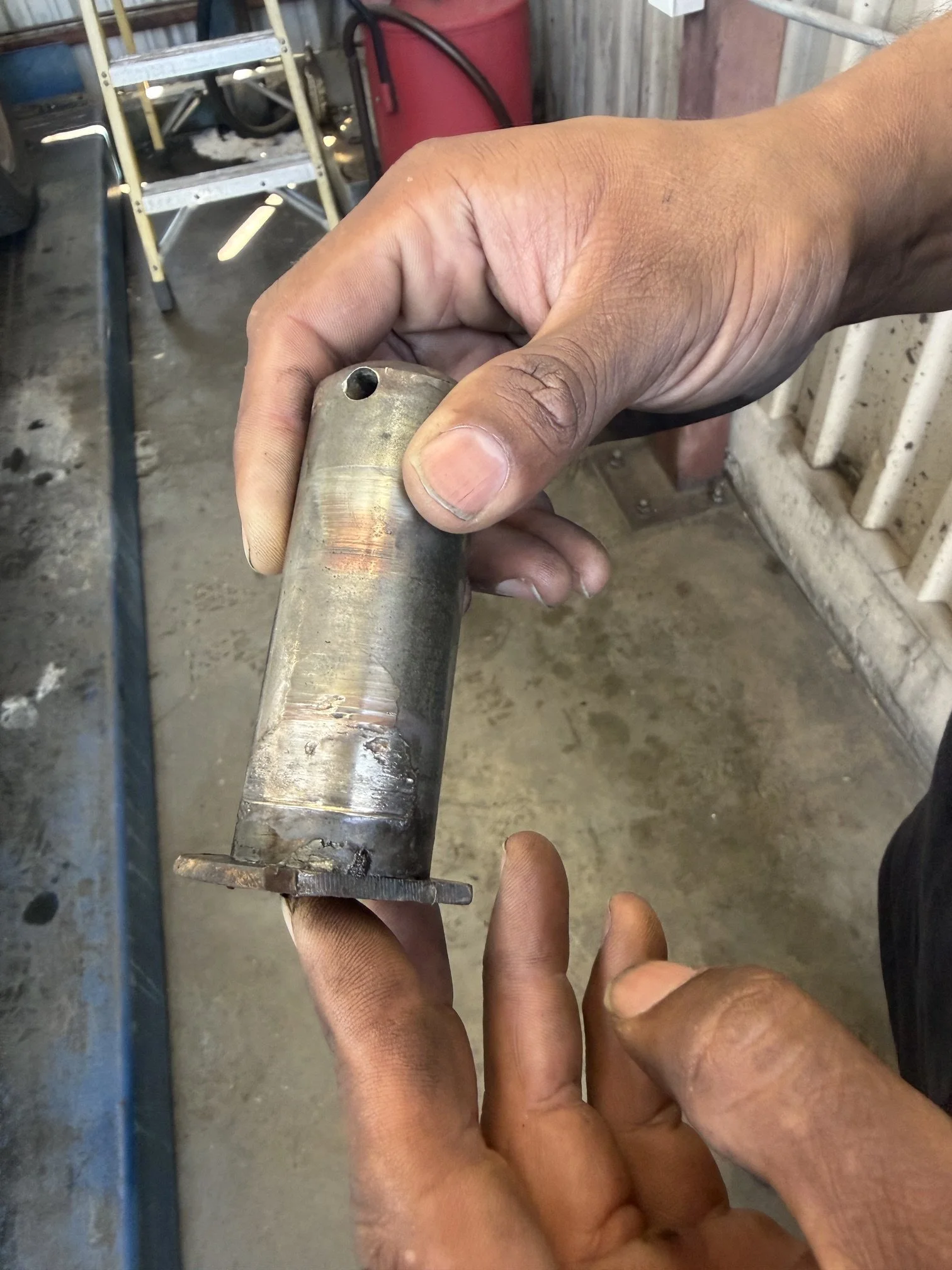 A person's hand holding a cylindrical metal object with a hole on top, in a workshop or garage setting.