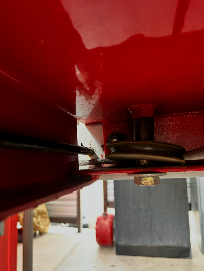 Close-up view of a pulley mechanism under a red-painted metal surface with visible bolts and screws, possibly part of a machine or equipment.