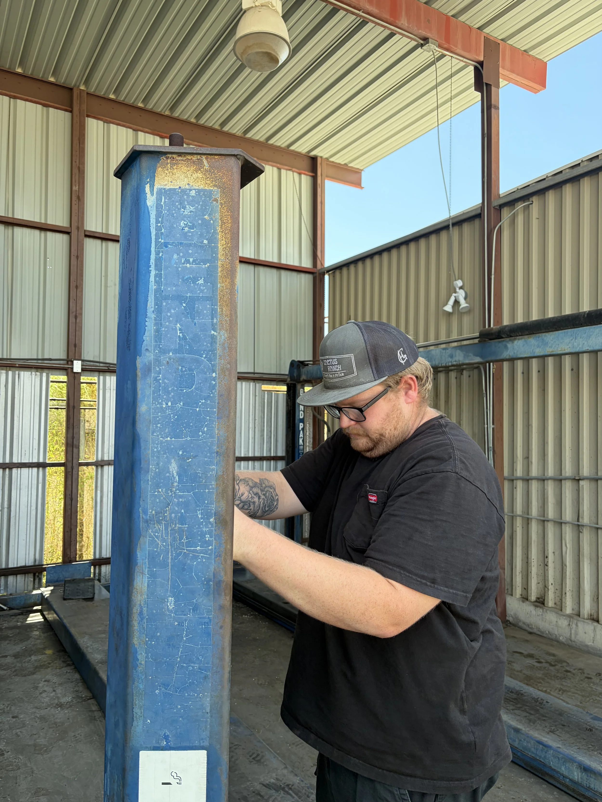 A man with glasses and a cap working outdoors under a metal roof, with a blue metal post in front of him and a fence in the background.