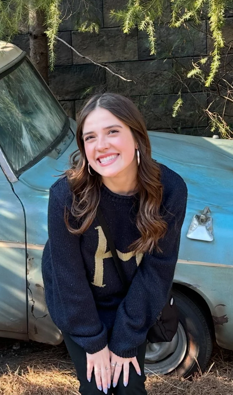 A young woman with brown hair and hoop earrings smiling while standing in front of a damaged light blue vintage car with a cracked windshield and dented body, outdoors with green foliage and a stone wall in the background.