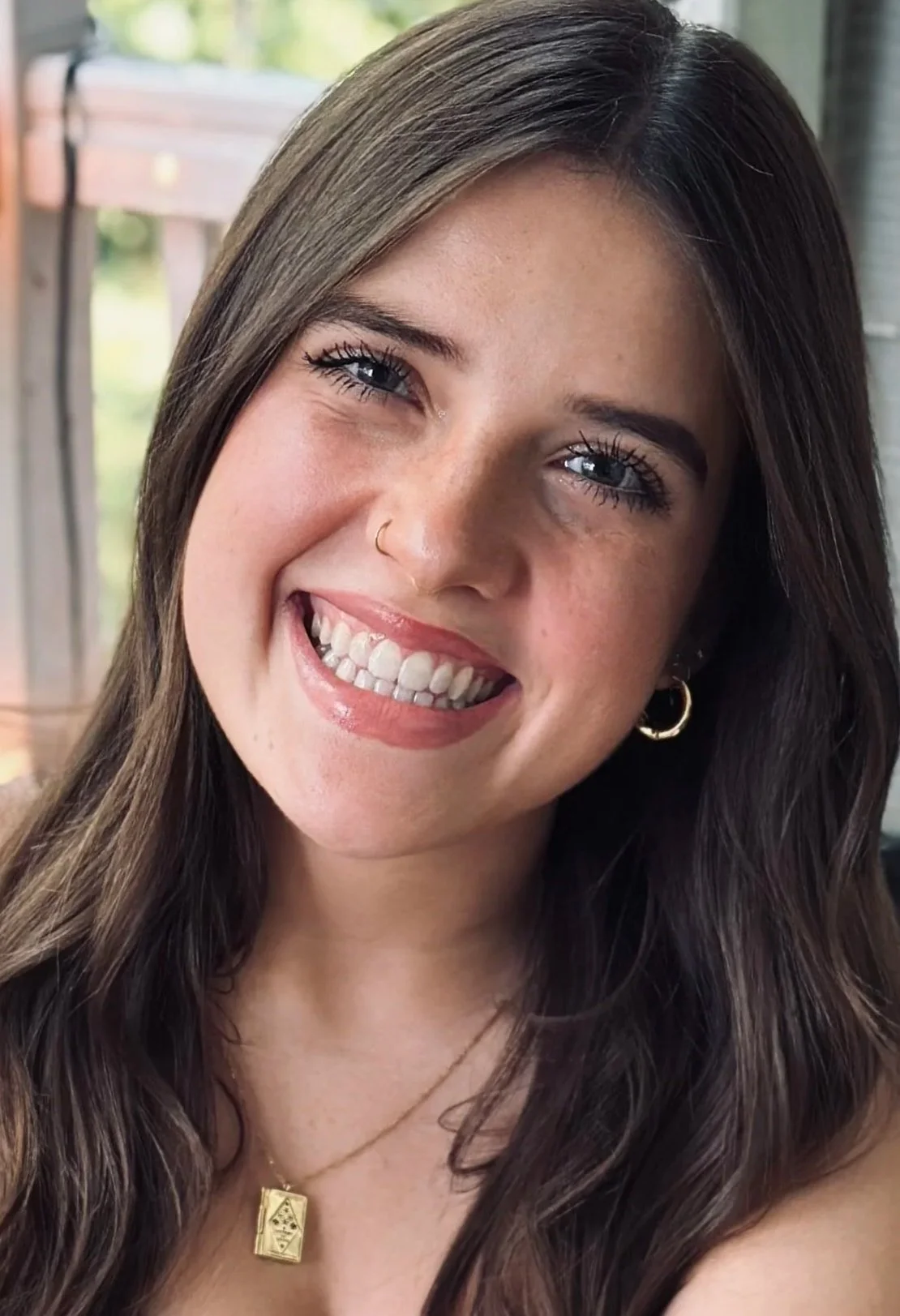 Close-up of a young woman smiling, showing her teeth, with blue eyes, dark brown hair, and wearing gold hoop earrings, a gold necklace with a rectangular pendant, and a nose ring.