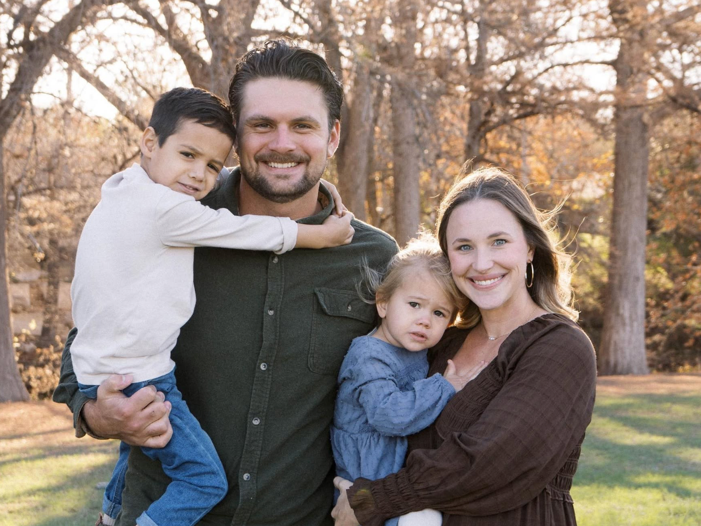 A smiling family of four outdoors in a park during autumn, with trees and fall foliage in the background. The father is holding a young boy on his side, while the mother is holding a young girl. All are looking at the camera.