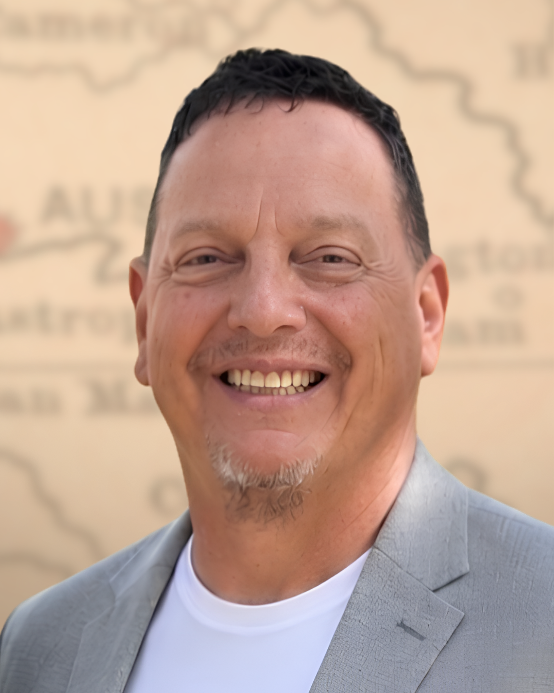 A smiling man with short black hair, a goatee, and wearing a light gray suit jacket over a white shirt, standing in front of a blurred map background.