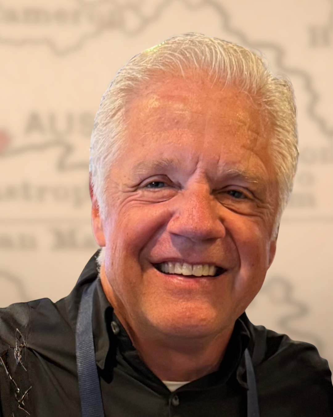 Smiling older man with light blonde hair, wearing a black shirt, in front of a blurred background with indistinct writing.
