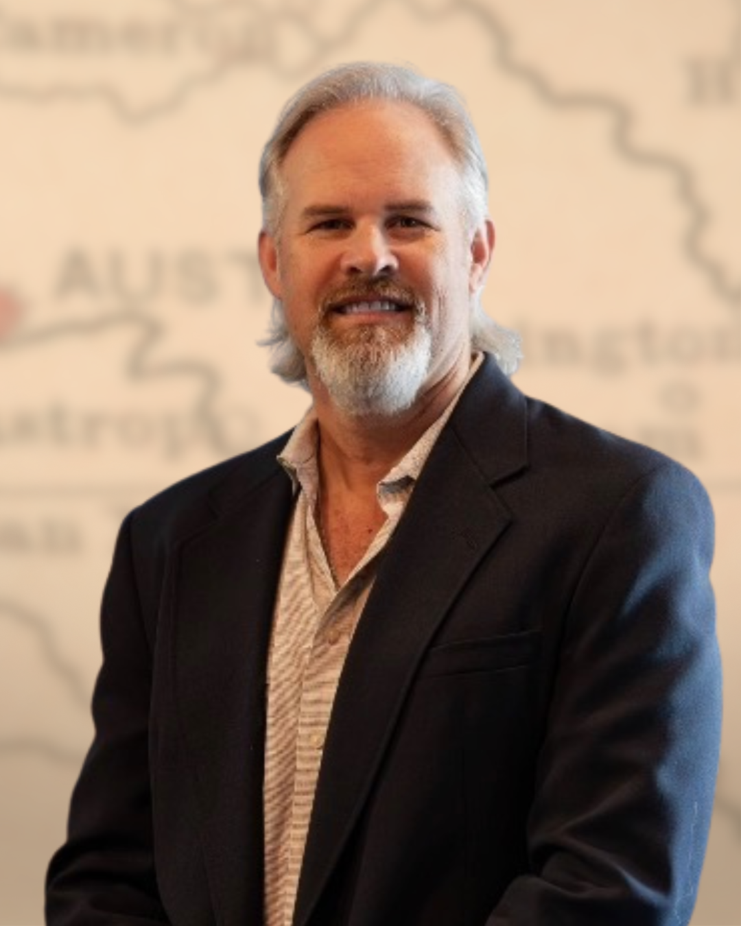 A middle-aged man with light gray hair and a beard, wearing a dark blazer and a patterned shirt, smiling at the camera with a blurred background that appears to be a map.