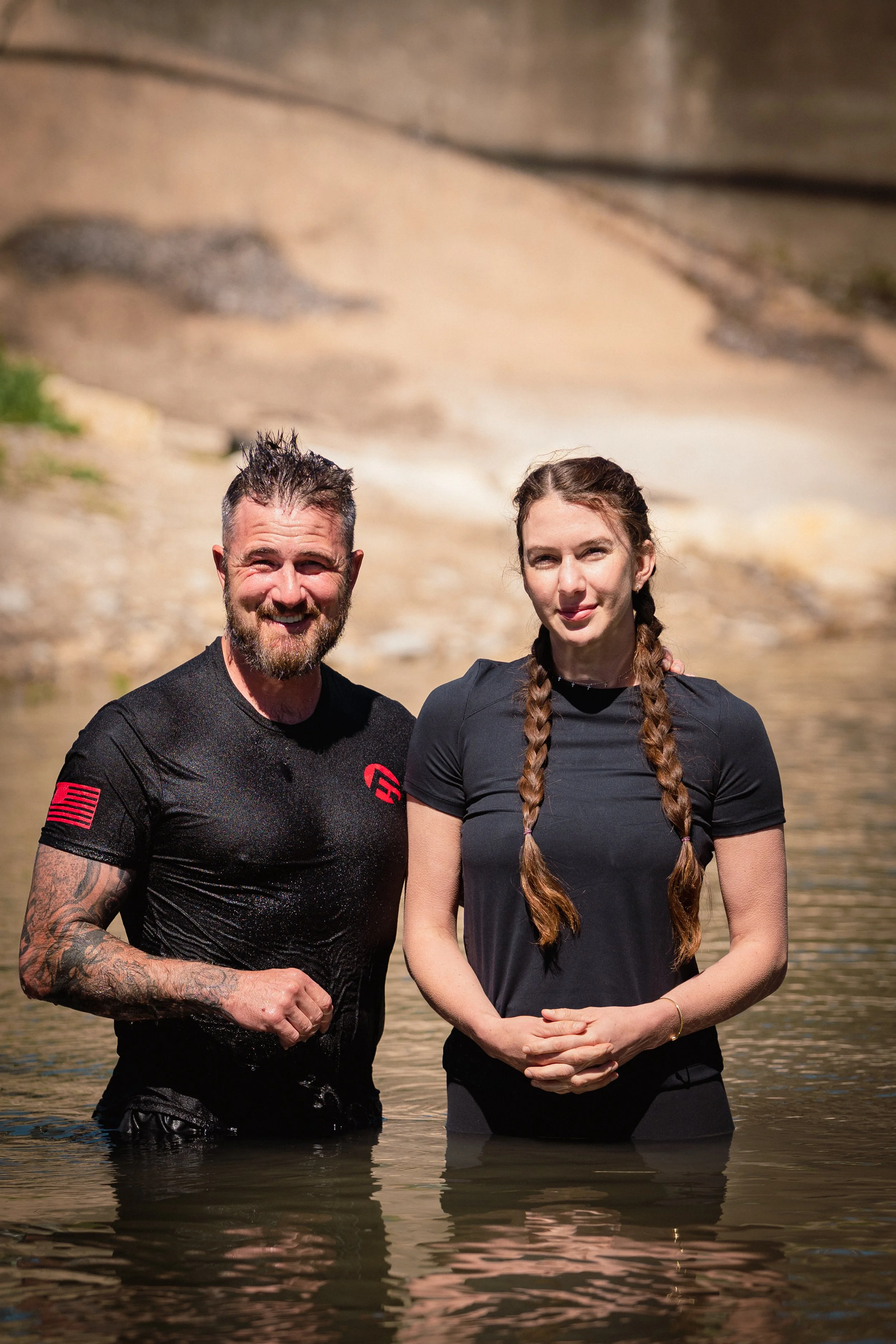 A man and a woman standing in a body of water, with a sandy and rocky background. The man has short, wet hair, a beard, and tattoos on his arm. The woman has long brown hair in braids and is wearing a dark athletic shirt.