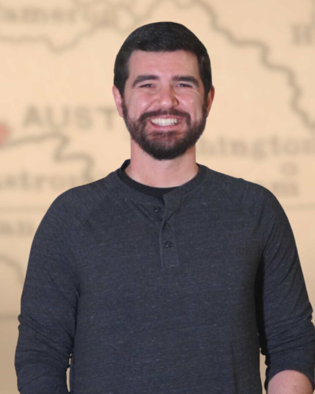 A smiling man with dark hair and a beard wearing a dark grey long-sleeve shirt, standing in front of a blurred background with a map and text.