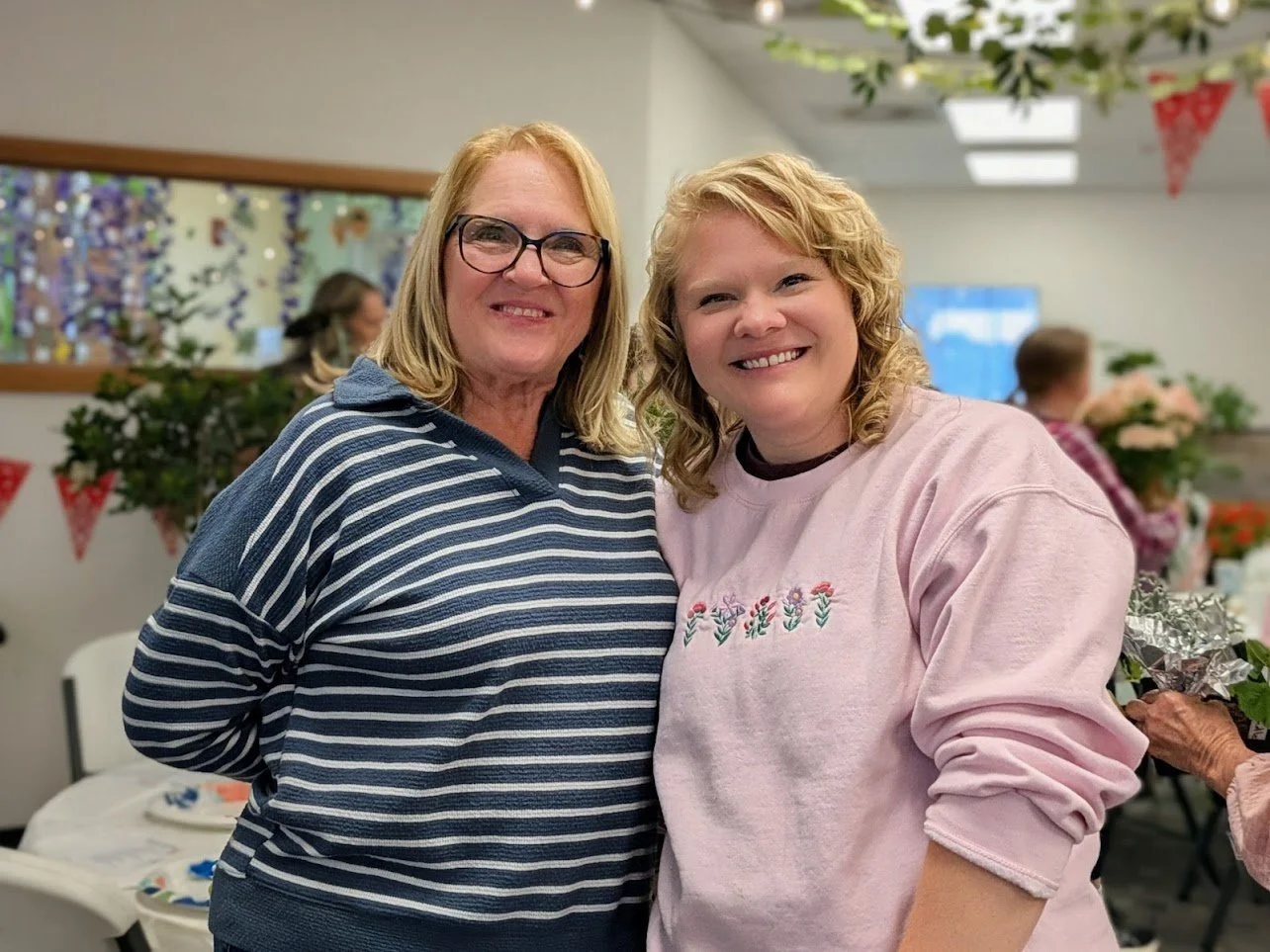 Two smiling women standing close together at a party or gathering, indoors with decorations and other people in the background.