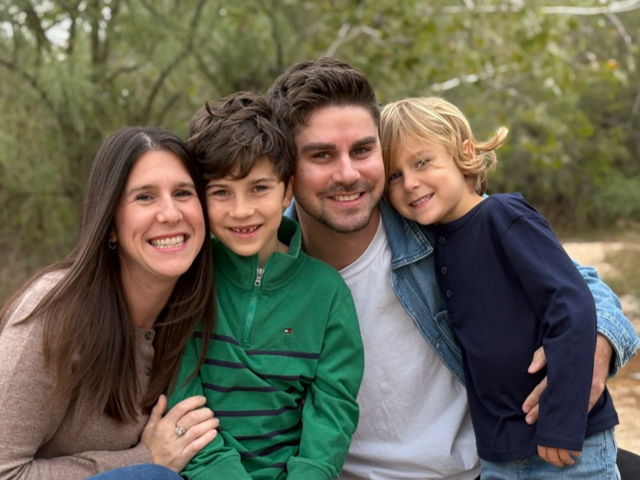A family of four smiling outdoors in a wooded area, with a woman, a teenage boy, a young man, and a young boy, all close together