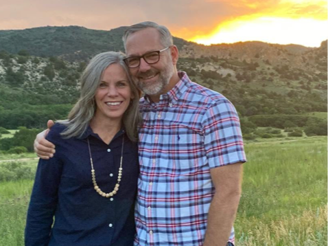 A smiling couple standing outdoors at sunset, embracing each other, with hills and a green field in the background.