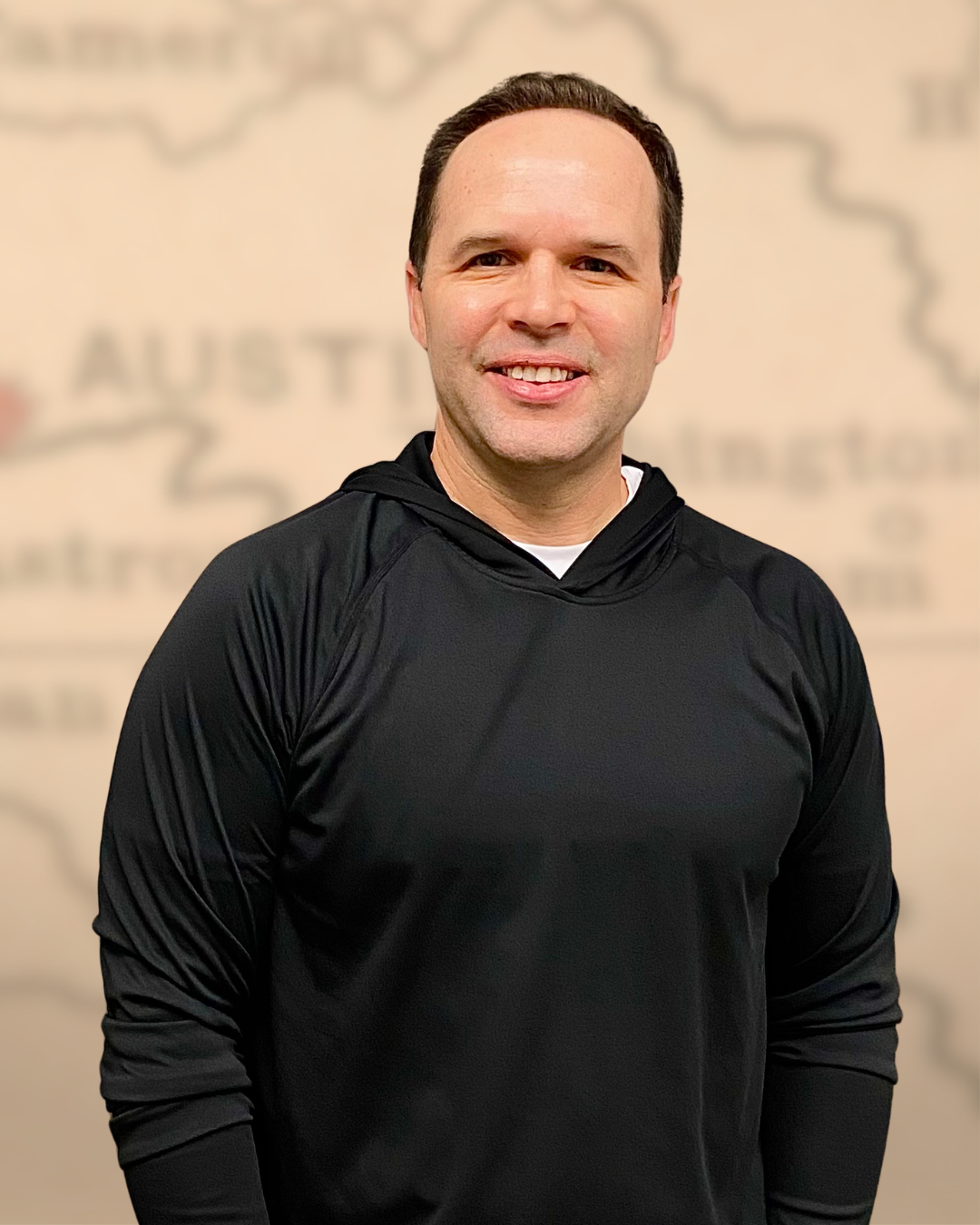 A smiling man with short brown hair and a fair complexion wearing a black athletic hoodie, standing in front of a blurred background with the word 'AUSTIN' visible.