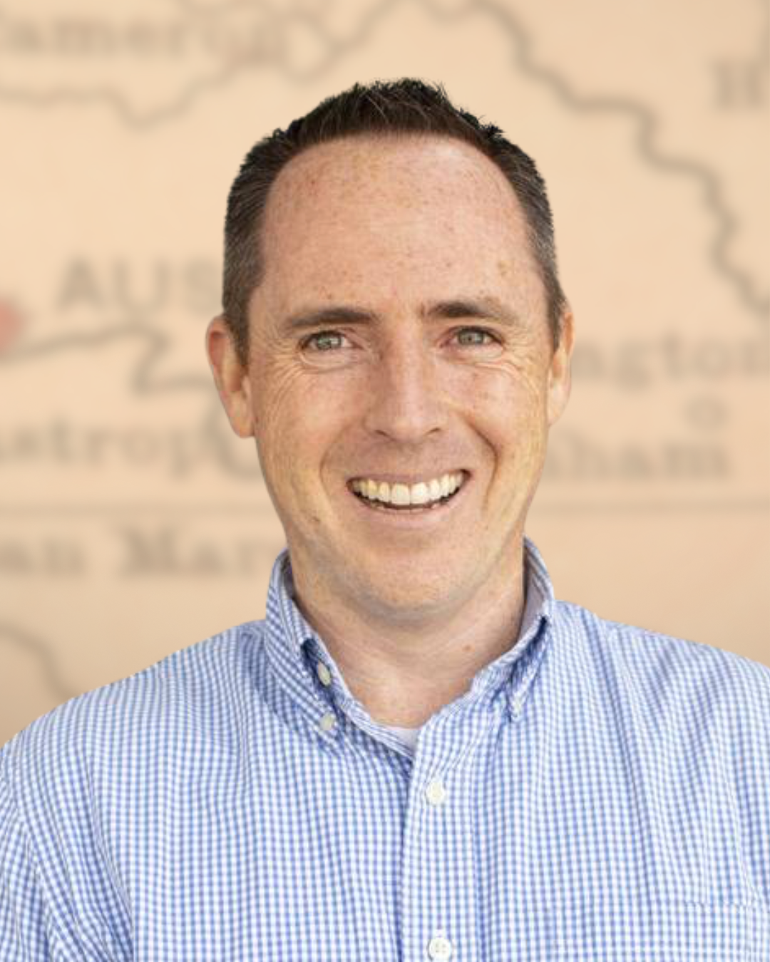 A smiling man with short dark hair wearing a blue and white checkered shirt, standing outdoors in front of a blurred background with some text.