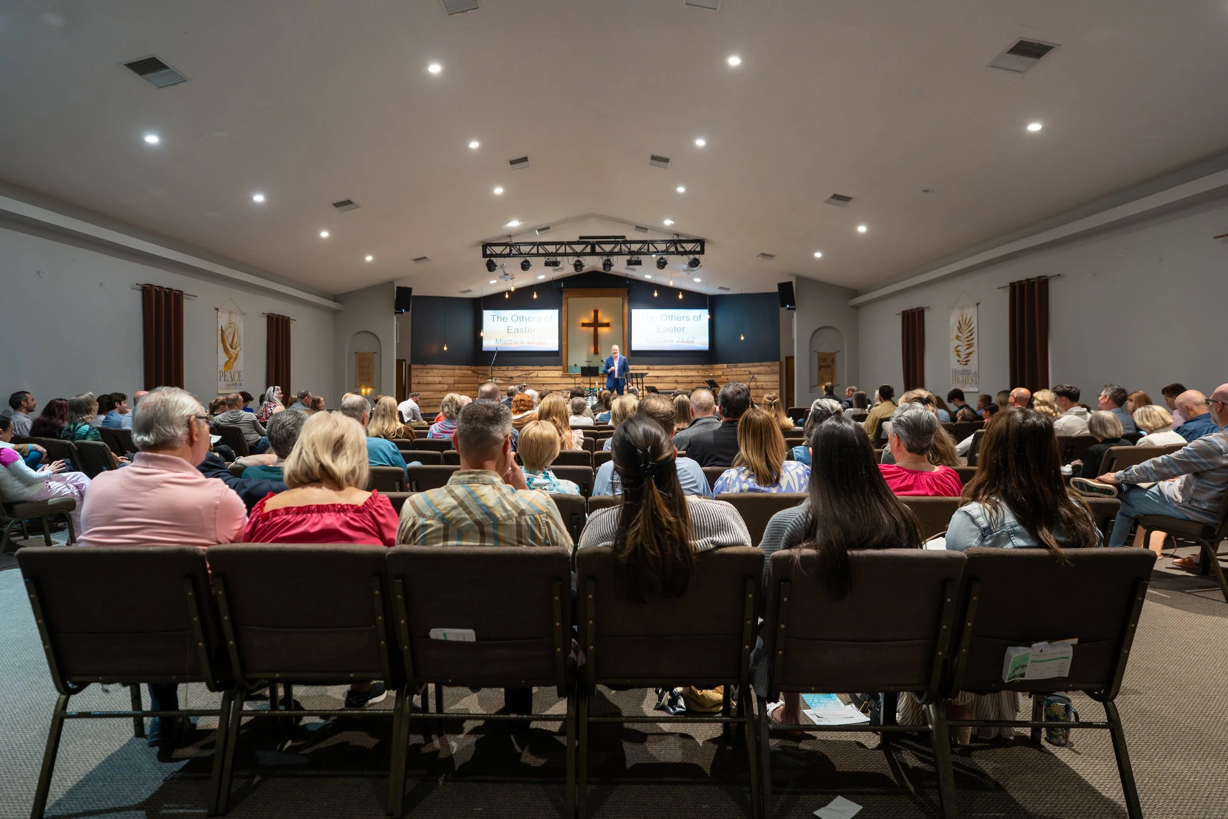 A church service with a preacher standing at the front in front of a cross, speaking to a congregation seated in pews; two screens behind him display sermon titles, and the church interior includes banners, curtains, and lighting.