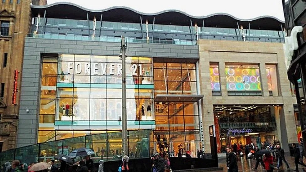 Multistory shopping mall with a large Foreve 21 sign on glass front and colorful window display, people walking in the rainy weather, some holding umbrellas.