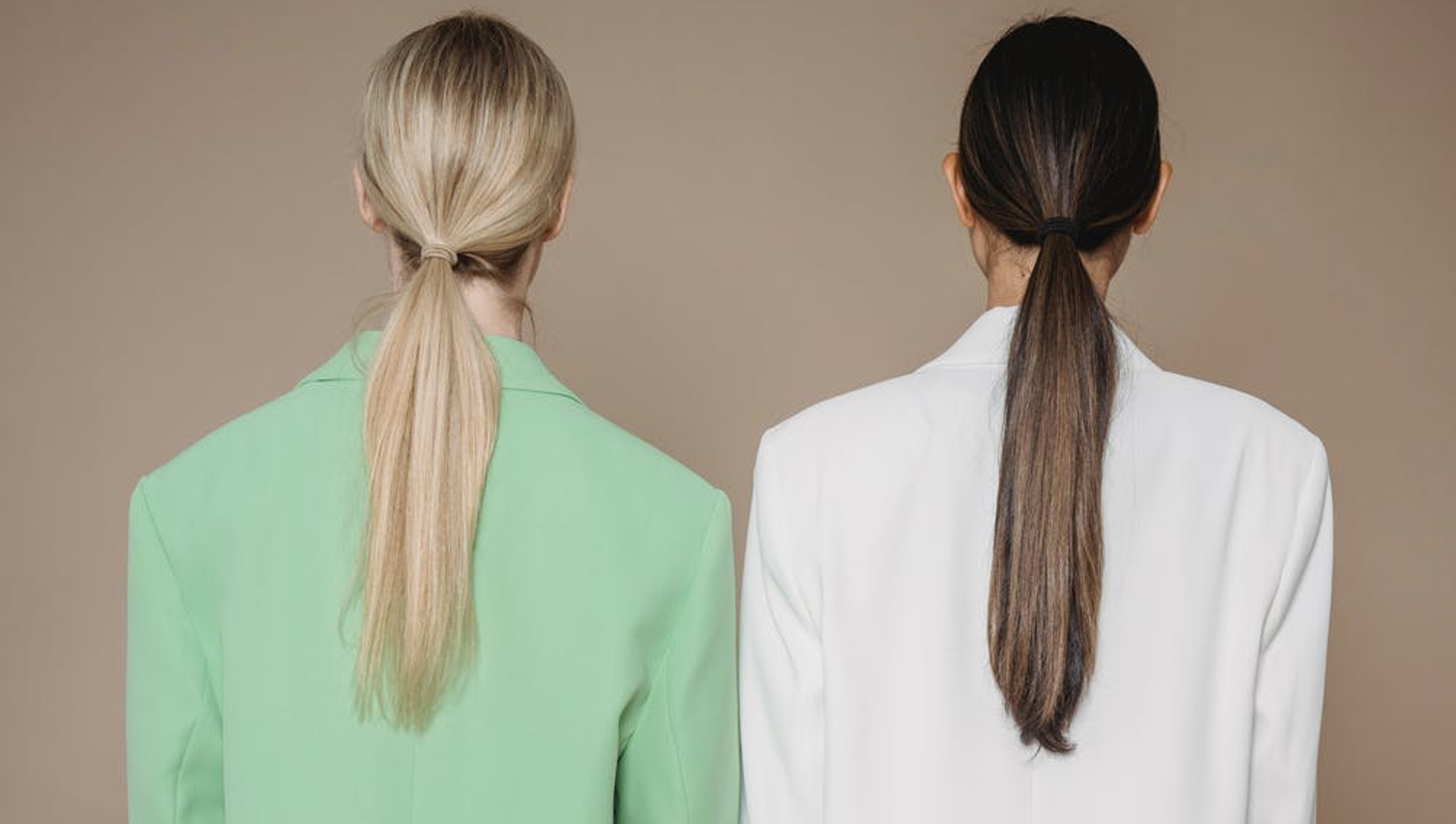 Two women with different hair colors and styles, seen from behind, wearing contrasting blazers, standing against a neutral background.