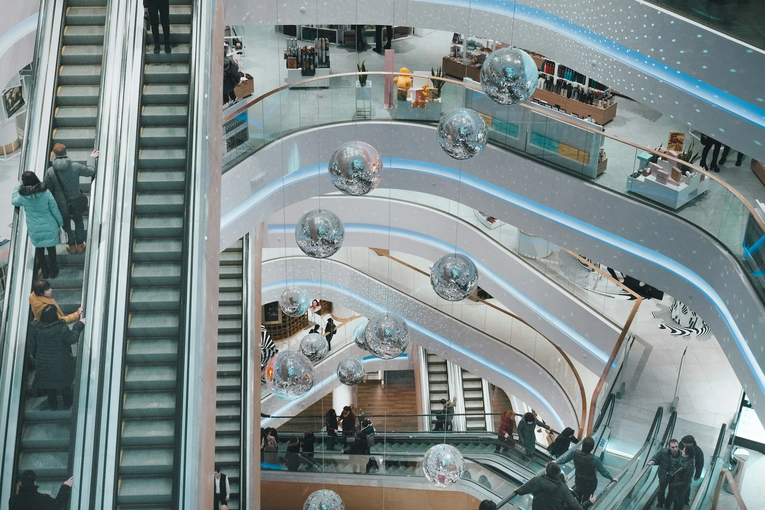 Multiple floors of a shopping mall seen from above, with escalators, shoppers, and decorative hanging disco balls.