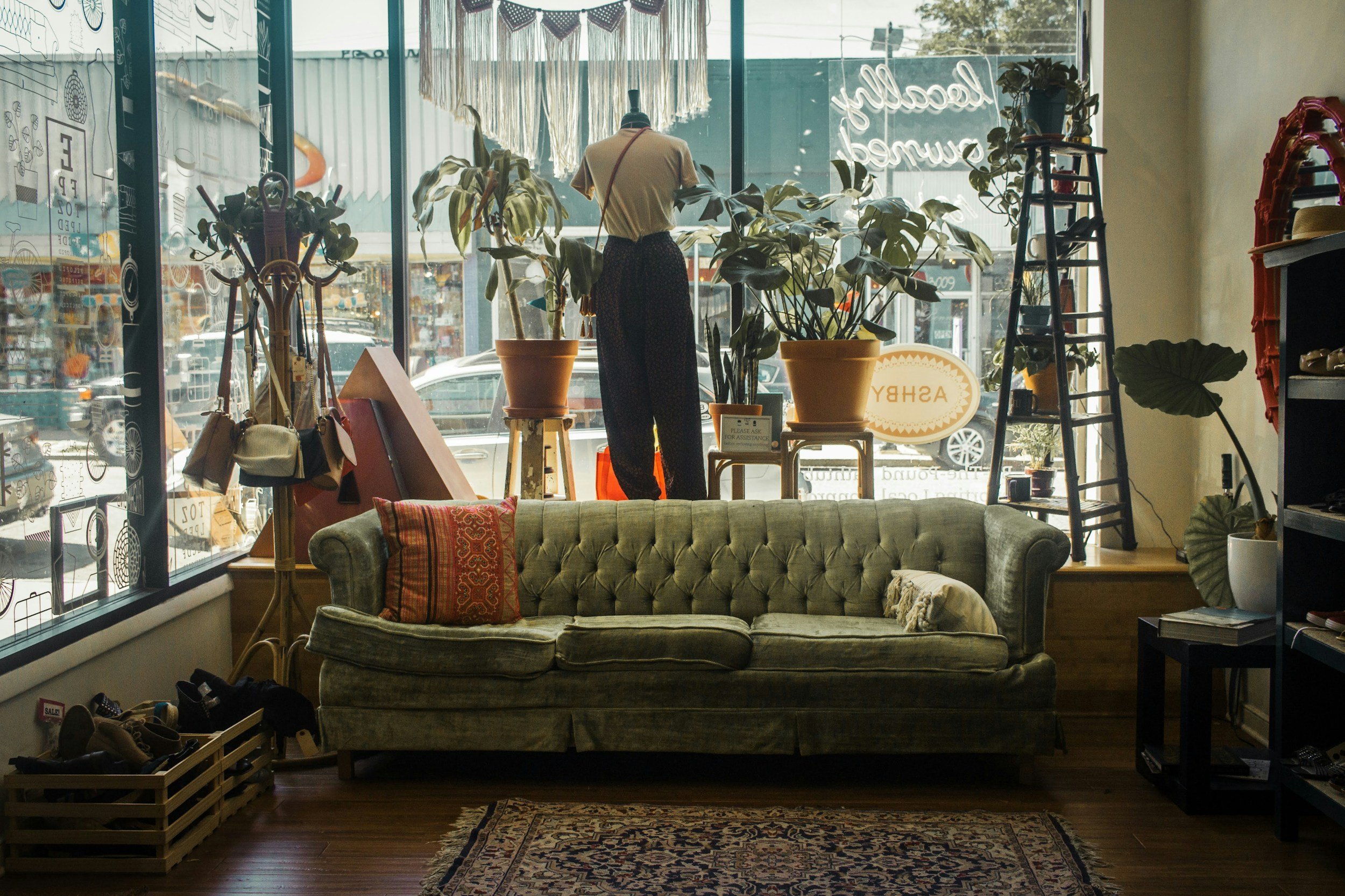 Inside a retail store showing a green vintage sofa with cushions, plants near the large front windows, and racks of shoes along with other furniture pieces. The store has a cozy, eclectic decor with natural light coming through the windows.