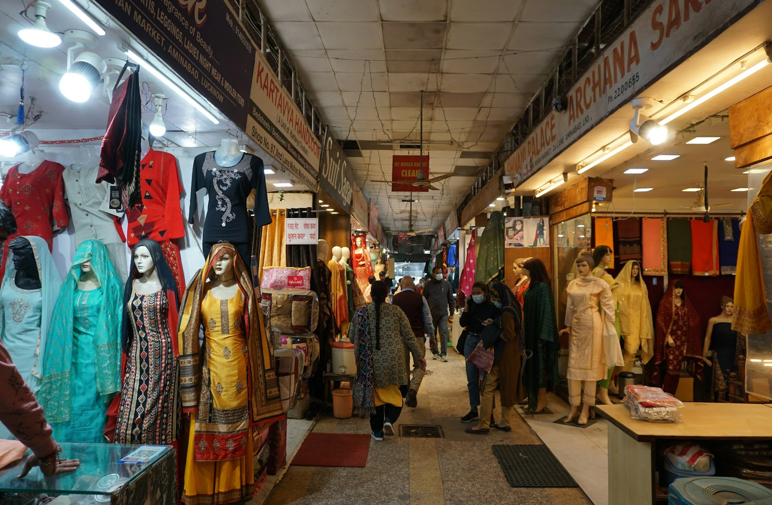 Indoor market with clothing stalls displaying traditional Indian dresses on mannequins; shoppers navigating through the market; overhead signs advertising different stalls and products.