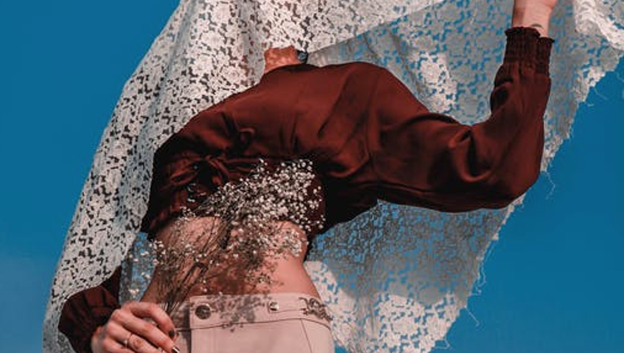 A fashion model's midsection with one hand on her hip, wearing a white lace top, maroon velvet jacket, a pink belt, and a floral skirt, against a plain blue background.